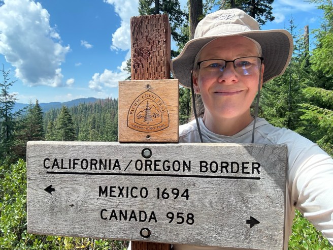 Me, standing behind the sign marking the border between California and Oregon. The sign has an arrow pointing south and reads "Mexico 1694"; below that is an arrow pointing north and the words "Canada 958."