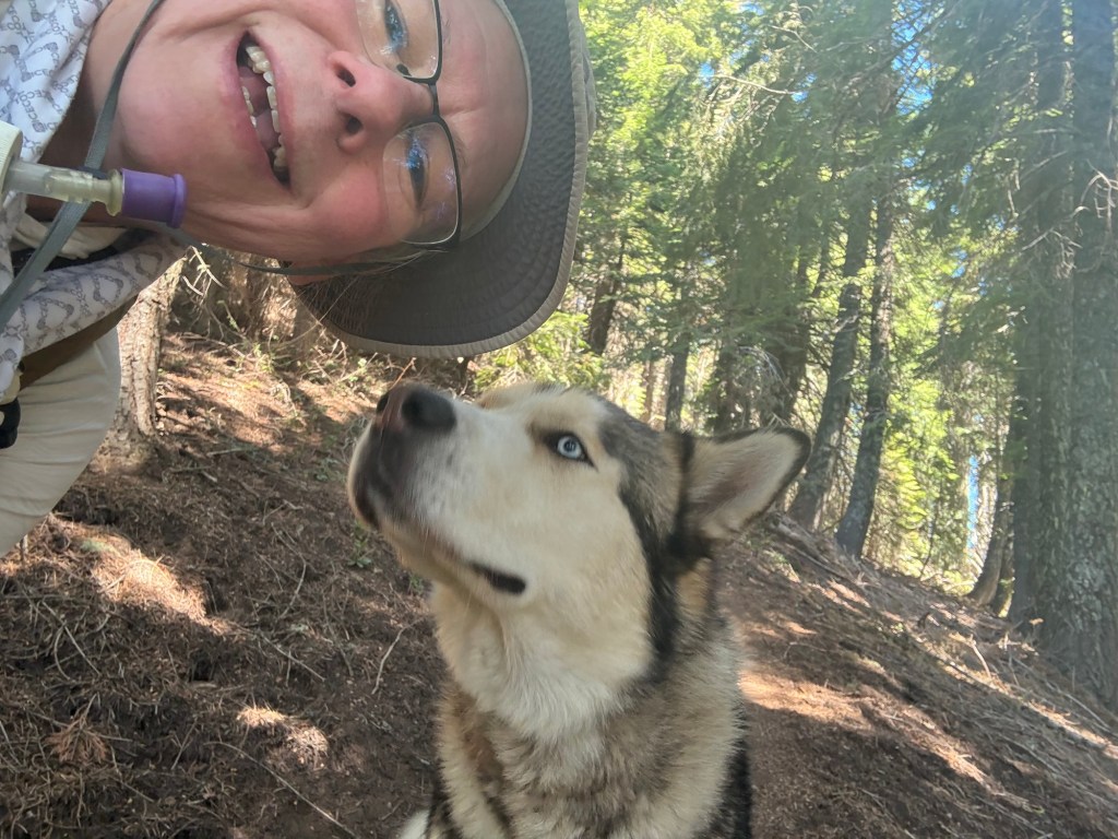 Selfie of me with a husky. I am looking at the camera, smiling. The dog is looking up at my face as if he is sniffing me or is otherwise curious about me.