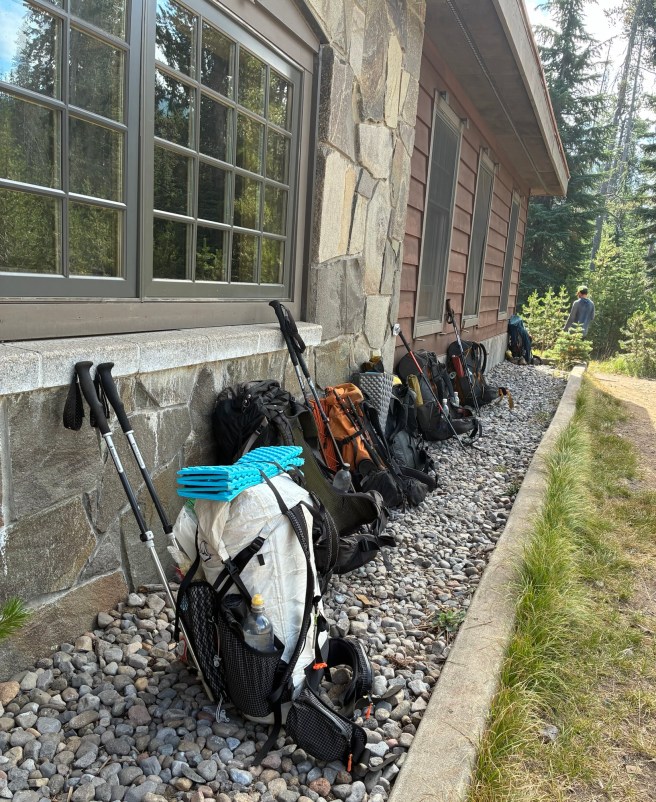 Photo of the exterior of a building at Mazama Village. There are a half-dozen backpacks lined up, leaning against the outside of the building.