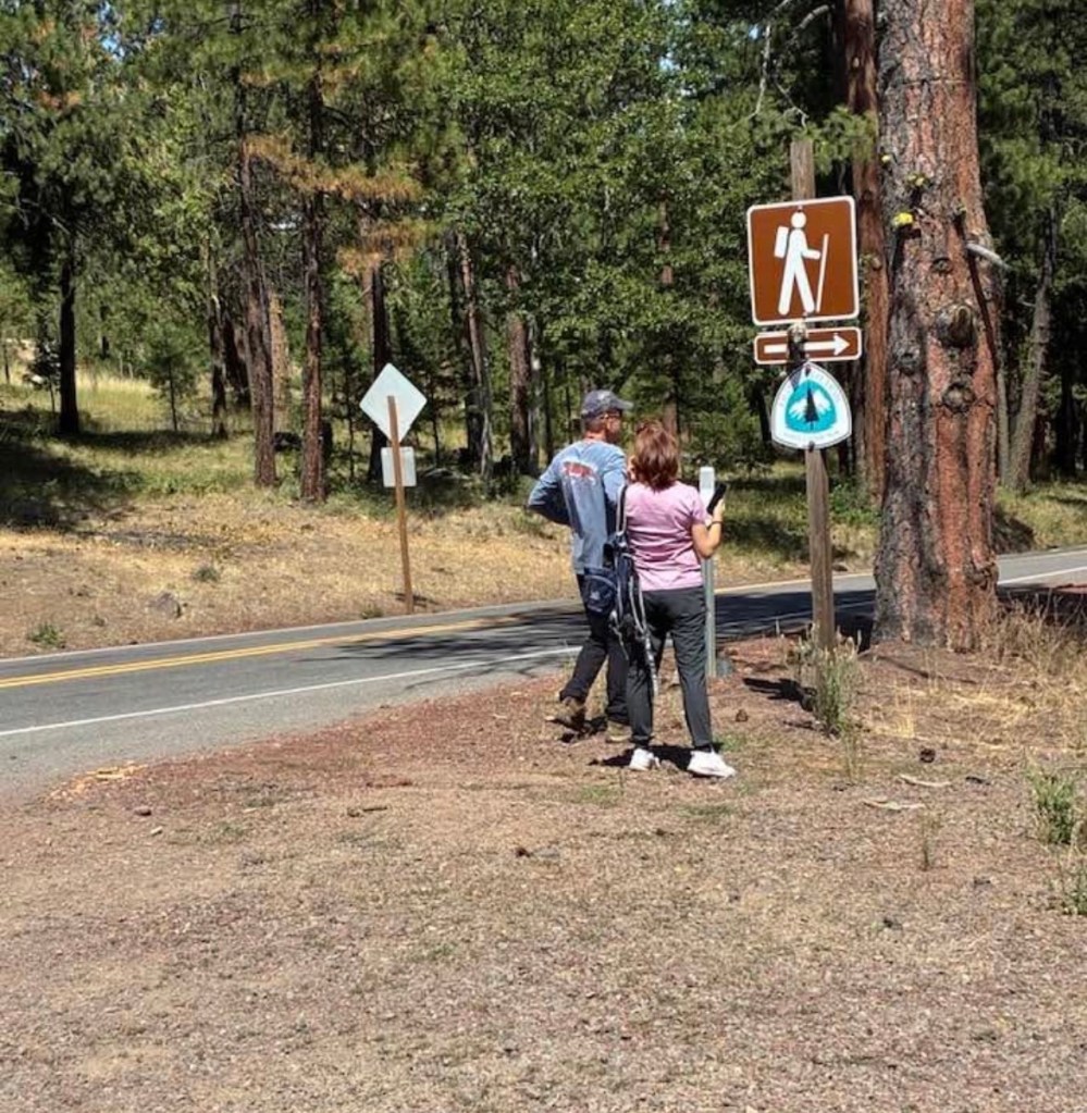 Two people, not hikers, standing in front of a large PCT sign and a sign indicating the direction of the trail. One of them appears to be taking a picture of the signs with her phone
